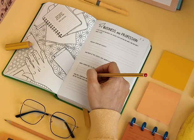 uma mesa amarela com detalhes laranja e uma agenda no centro com uma mão anatando algo a lápis